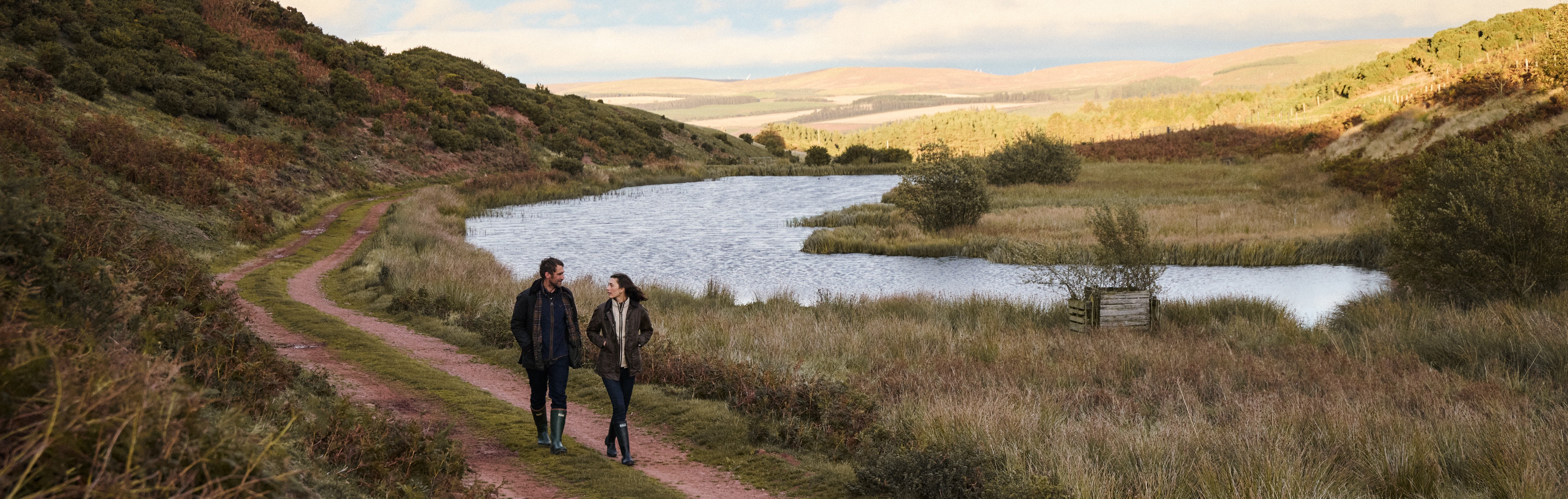 Twee wandelaars in Barbour jassen en laarzen tijdens een wandeling langs een meer in het Britse landschap.